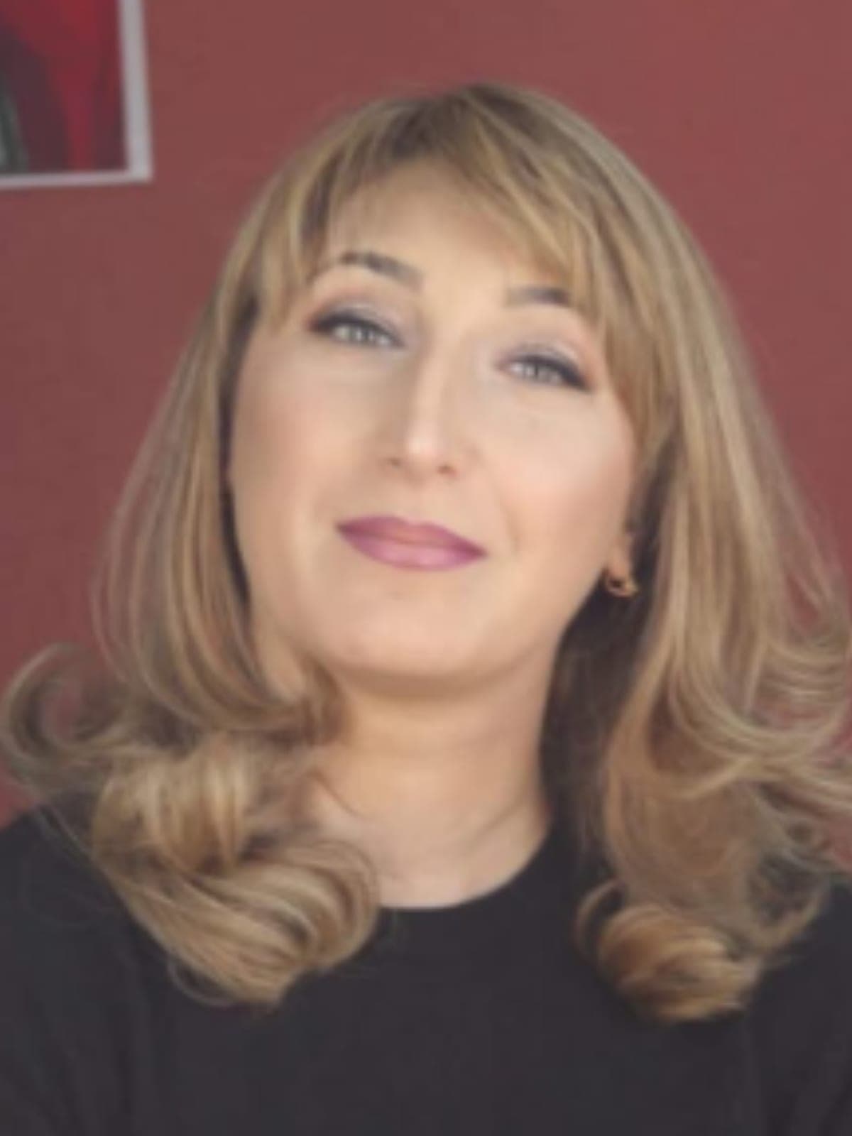 Teona: layered caramel-blonde hair in a soft curled blowout, wearing a black top against a deep red wall